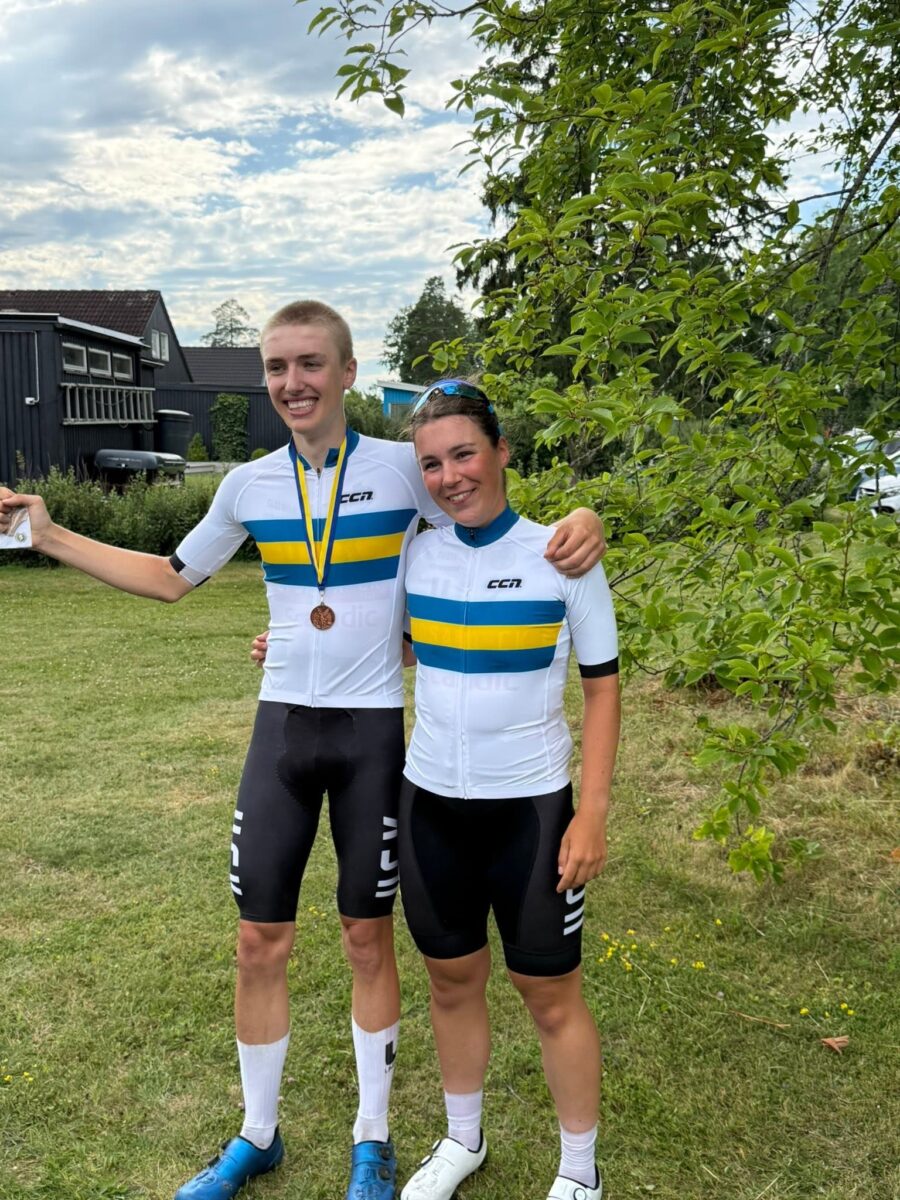 Two cyclists in matching white, blue, and yellow jerseys pose with arms around each other on a grassy field, one wearing a bronze medal.