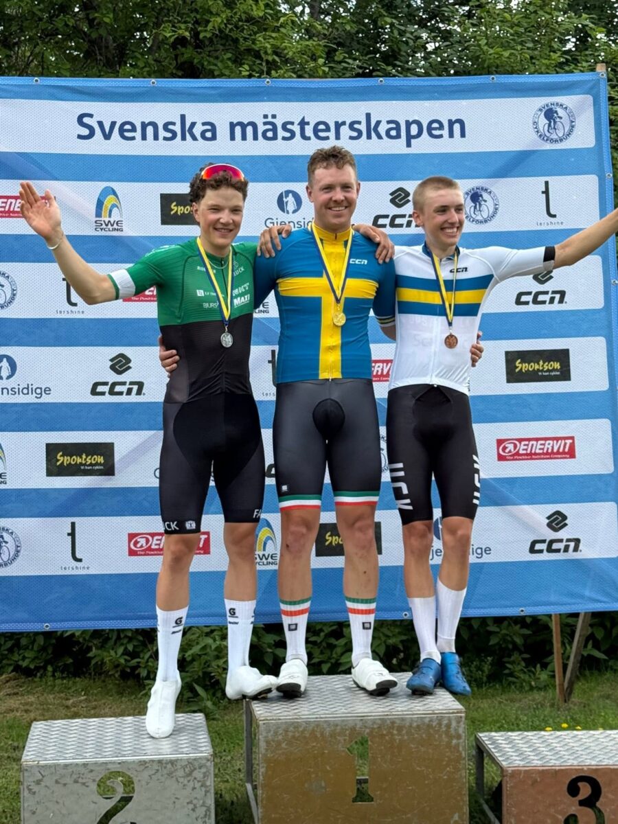 2 | Ski&Bike Three male cyclists stand on a podium with medals after a Svenska mästerskapen race, center in blue and yellow jersey as first place, flanked by green and white jerseys, cheering fans backdrop with sponsor banners.