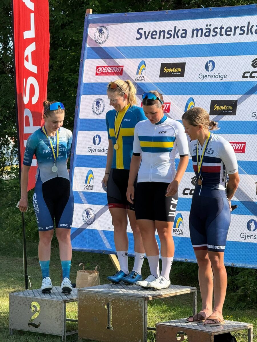 3 | Ski&Bike Four female cyclists on a winners' podium with medals, standing on platforms labeled 1, 2 and 3, in team kits with a sponsor banner behind them.